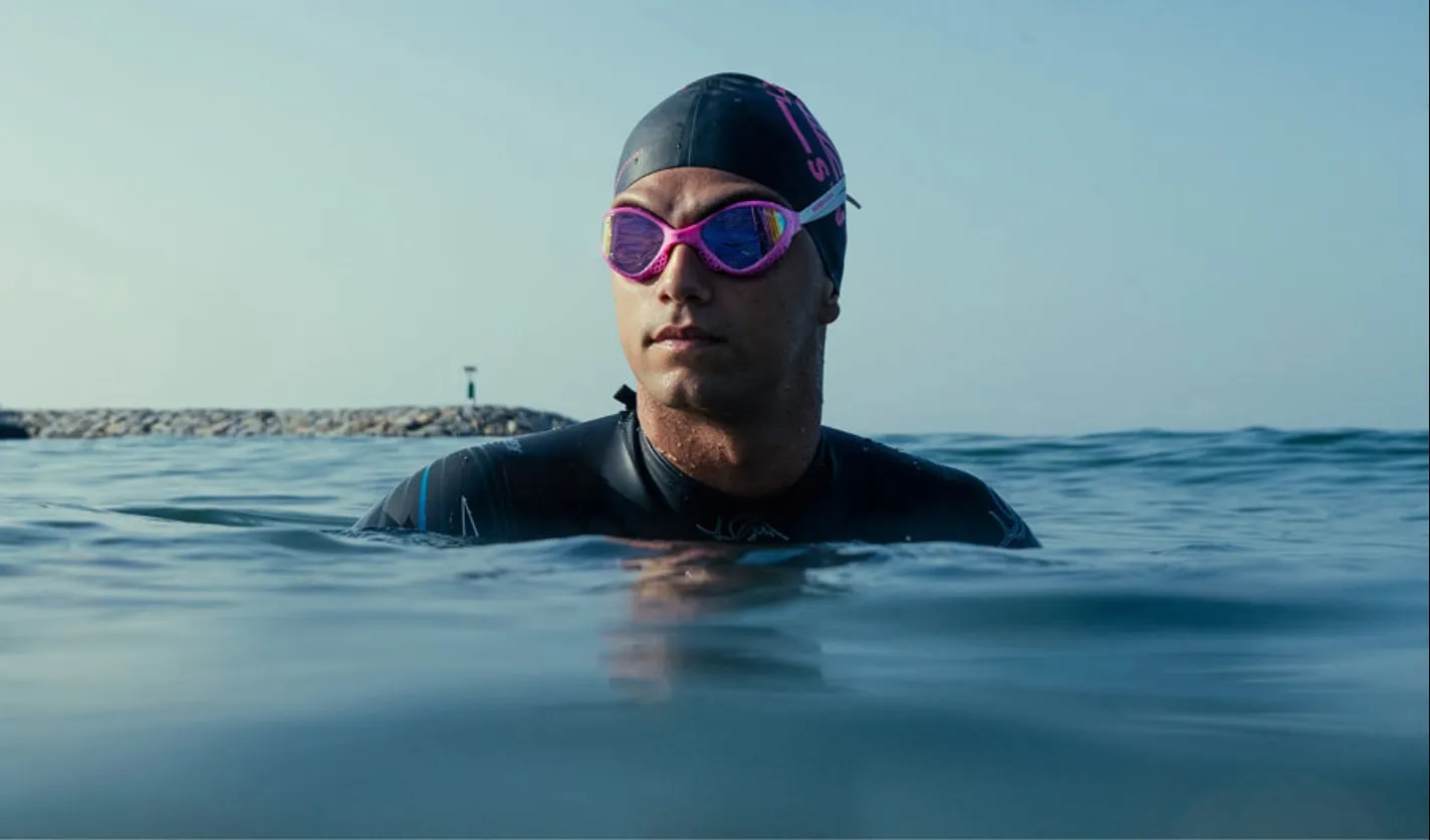 An open-water swimmer in a black wetsuit and pink mirrored Buddyswim goggles, head and shoulders above calm sea